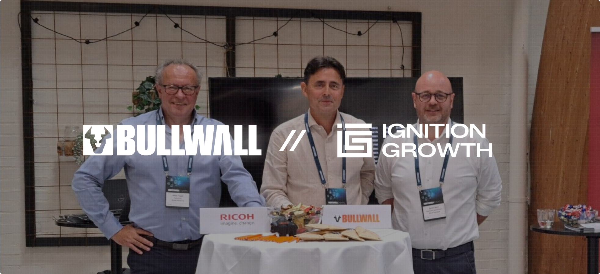 Three men standing behind a round table with snacks, displaying Bullwall and Ignition Growth logos.