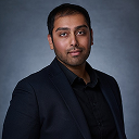 Portrait of a man wearing a black suit and shirt against a dark gray background.