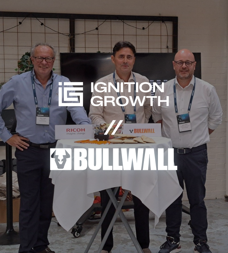 Three men wearing badges stand behind a round table with a white tablecloth and food, with logos for Ignition Growth, Ricoh, and Bullwall overlaid on the image.