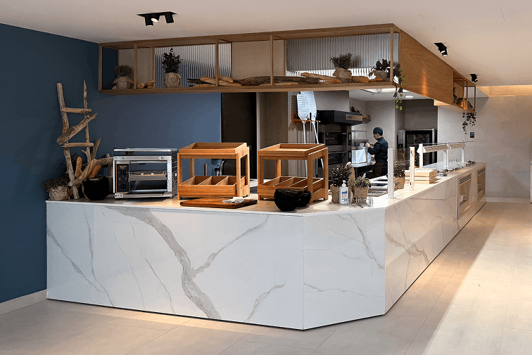 Modern kitchen counter with white marble finish, wooden display boxes, small appliances, and a person preparing food in the background.