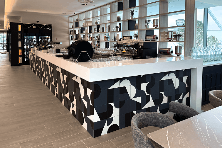 Modern cafe interior with a geometric patterned counter, coffee machine, and shelving with decor and plants under natural light.