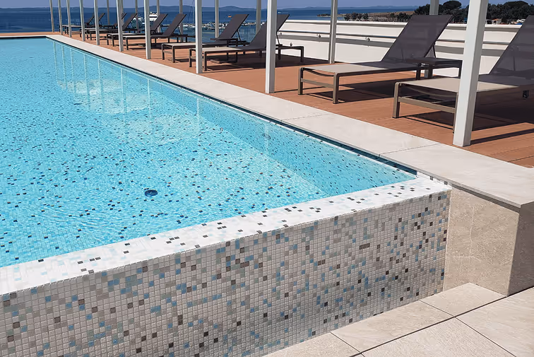 Infinity pool with light blue water and mosaic tile edge overlooking a calm sea and distant islands, with lounge chairs on a wooden deck.
