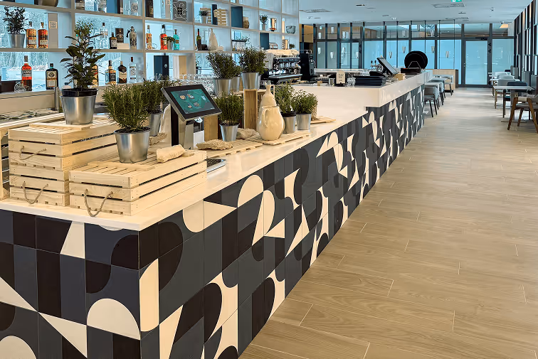 Modern bar counter with black and white geometric patterned front, decorated with potted plants and bottles on shelves behind.