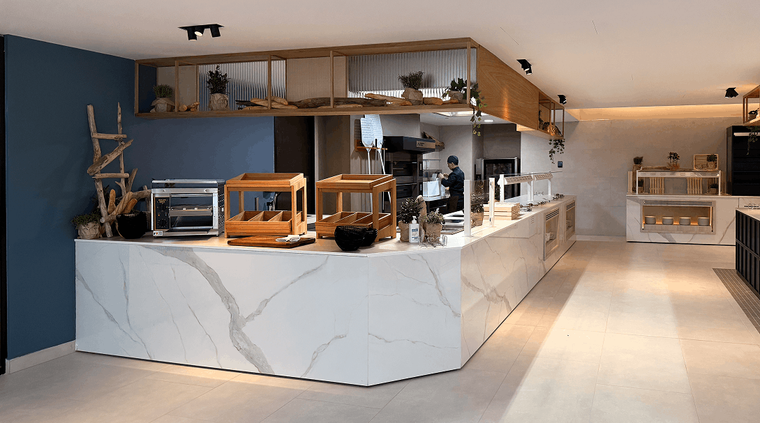 Modern bakery counter with marble facade, wooden display cases, bread oven, and a person preparing food in the background.