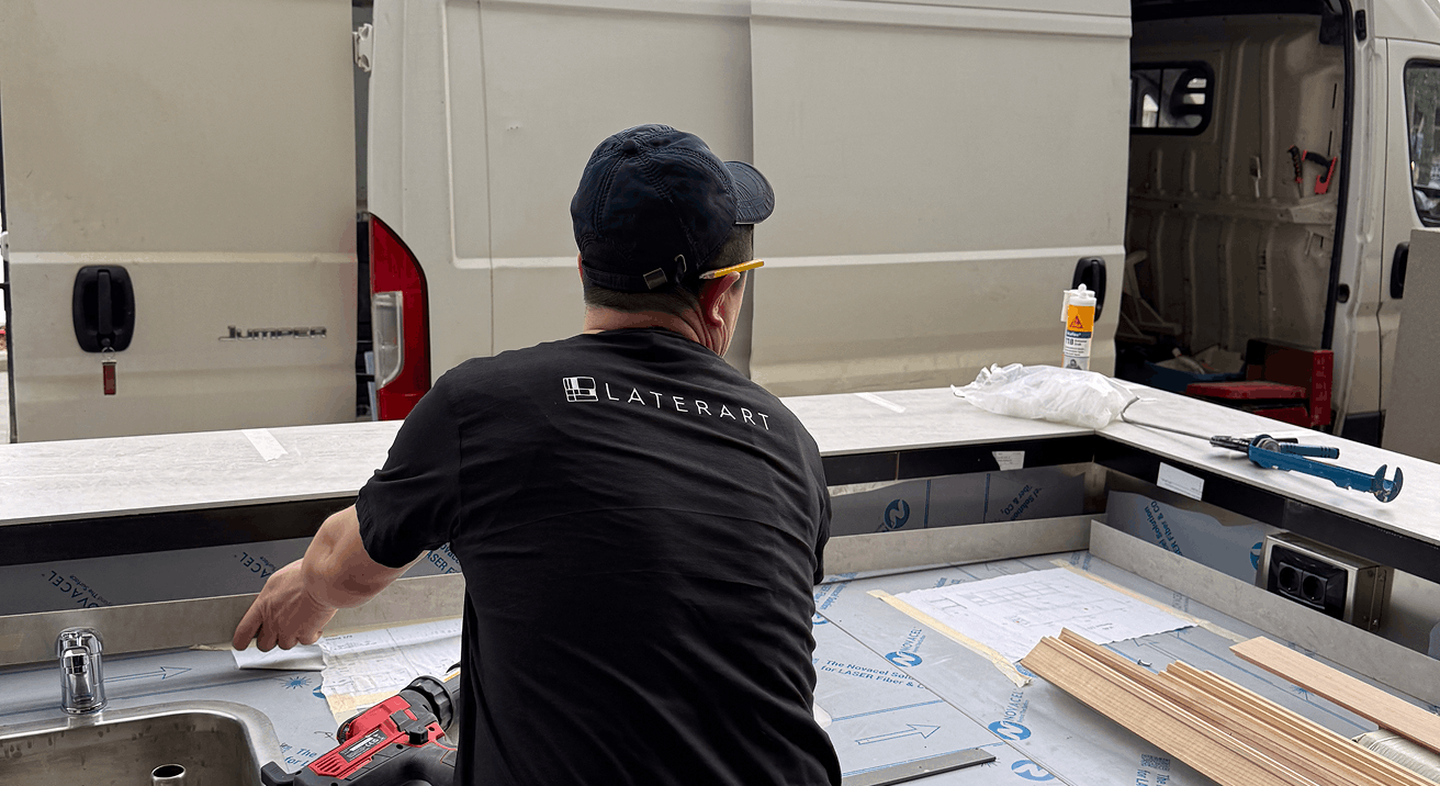 Man wearing a LaterArt shirt working on countertop installation inside a van with tools and wooden strips nearby.