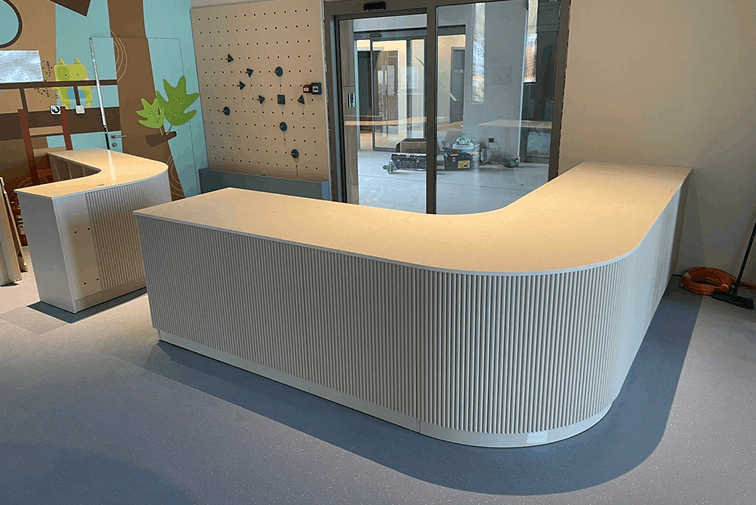 Modern, curved white reception desks in a room with blue flooring and a pegboard wall, near glass doors.