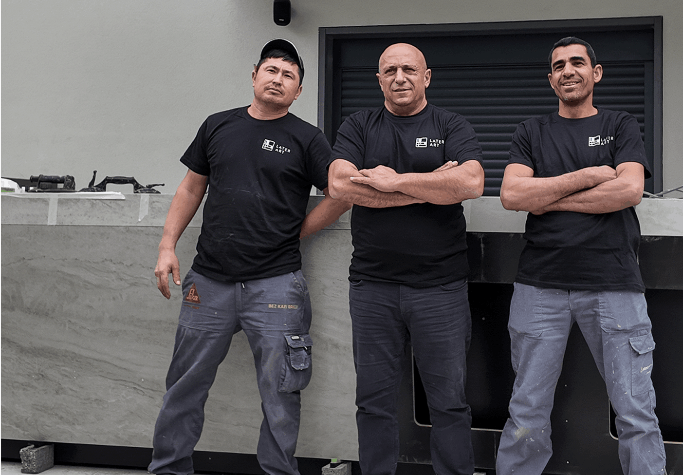 Three men wearing black Later Art t-shirts and work pants standing with arms crossed in front of a gray stone counter.