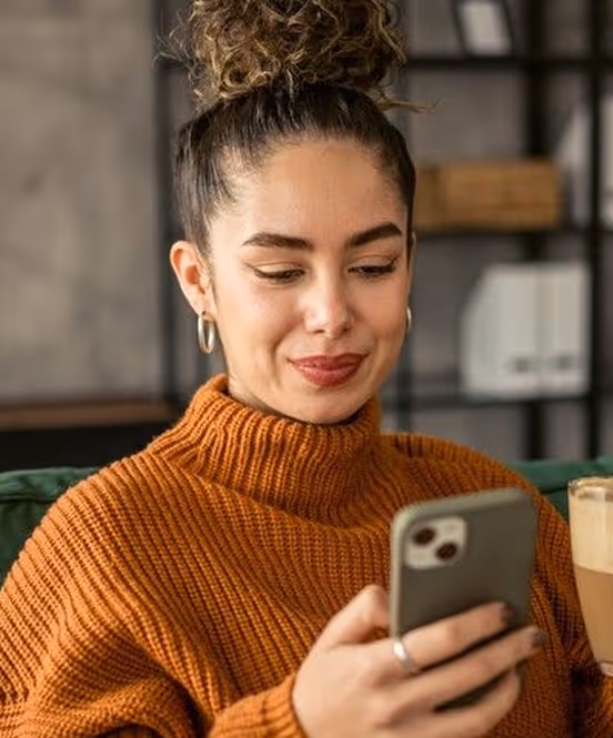 Young woman with curly hair in a bun wearing a brown sweater, looking at a smartphone while holding a glass of latte.