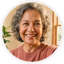 Smiling mature woman with curly gray hair wearing a pink shirt indoors near a window.