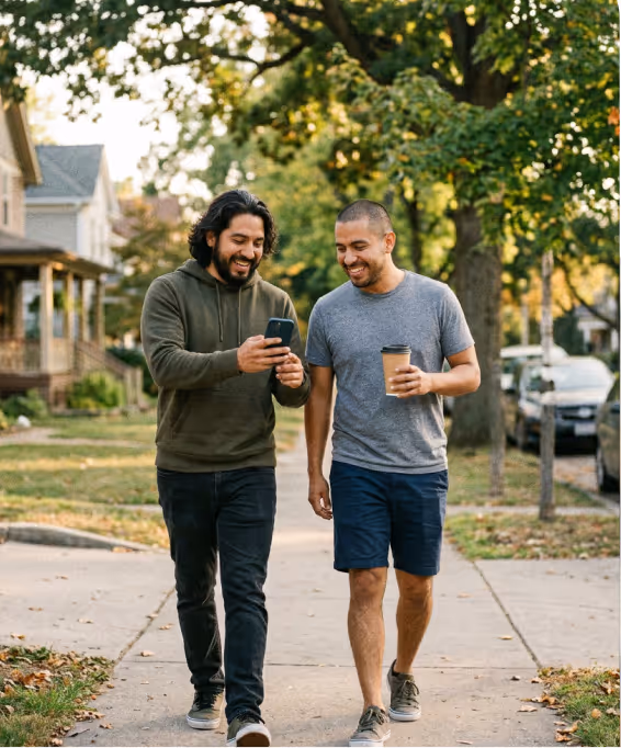 Two men walking on a sidewalk, one showing phone to the other who holds a coffee cup, smiling.