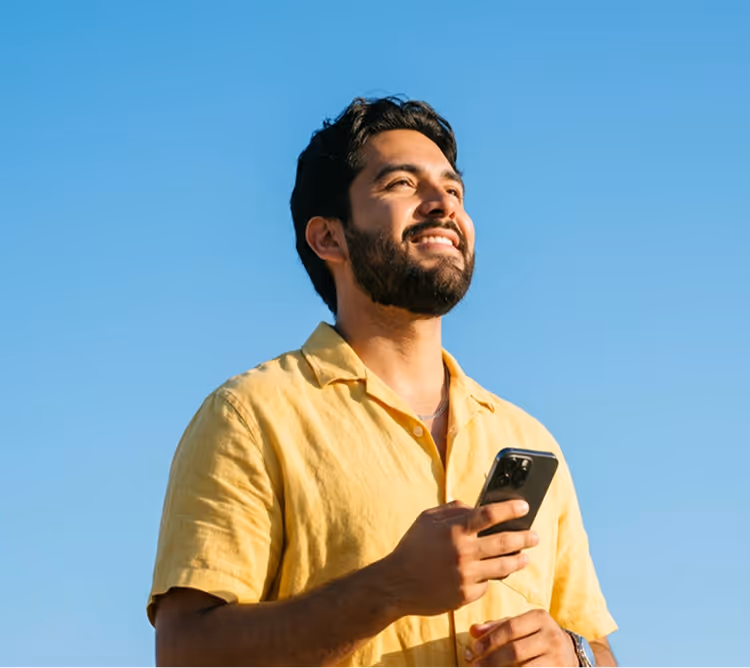 Smiling bearded man in yellow shirt holding smartphone under clear blue sky.