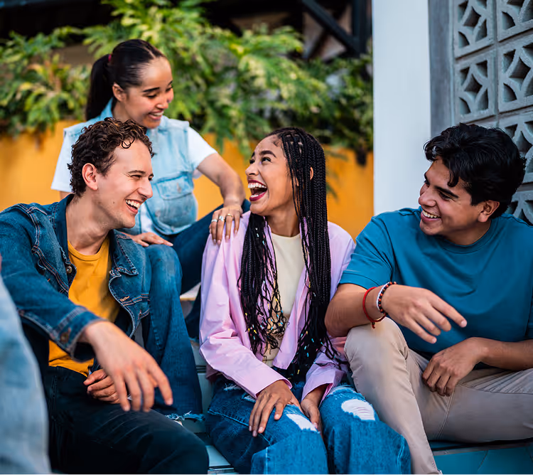 Four diverse friends sitting on stairs, smiling and laughing together outdoors.