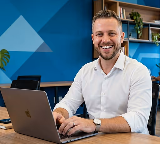 Smiling man in white shirt typing on laptop at office desk with blue wall and shelves behind him.