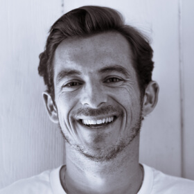 Smiling man with short dark hair and light facial stubble wearing a white shirt against a light background.