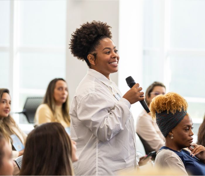 Smiling woman standing and speaking into a microphone in a seated audience at a meeting or conference.