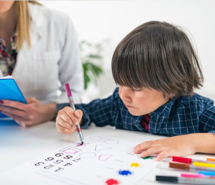 Child coloring shapes and numbers on a worksheet with markers while a woman in a white coat observes and takes notes.