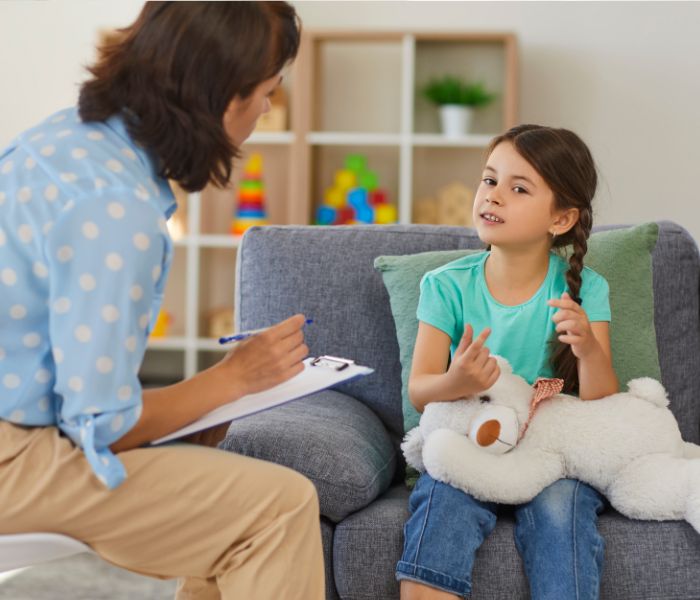 Young girl with braided hair sitting on a couch holding a white teddy bear, talking to a woman with a clipboard.
