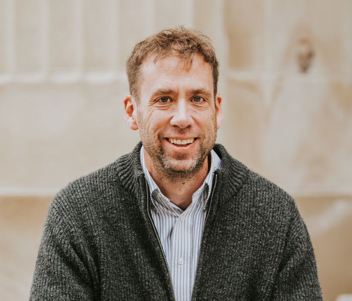 Smiling man with short curly hair wearing a striped shirt under a dark gray zip-up sweater, standing against a beige wall.