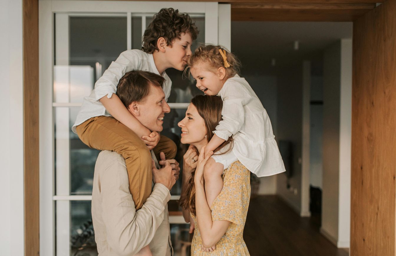 A smiling family of four inside a home, with father holding a boy on his shoulders and mother holding a girl on her shoulders as the children lean in close to their parents' faces.