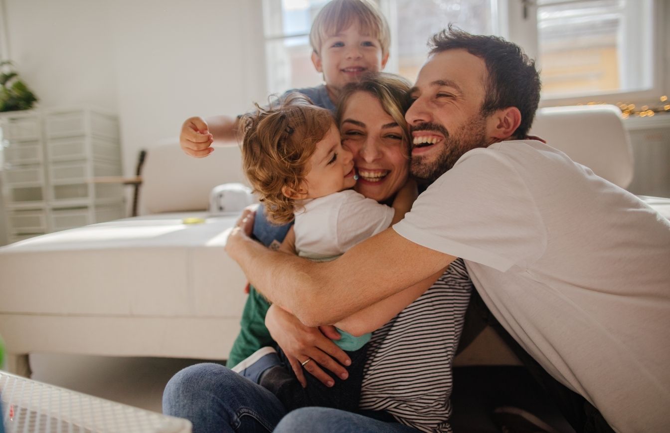 A joyful family of four hugging and smiling in a bright living room.
