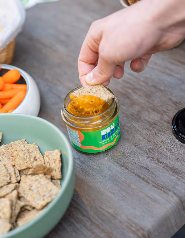 Une main trempe un cracker dans un petit pot de tartinade végétale à base de Carotte & Zaatar sur une table en bois, avec un bol de crackers et des carottes en arrière-plan.