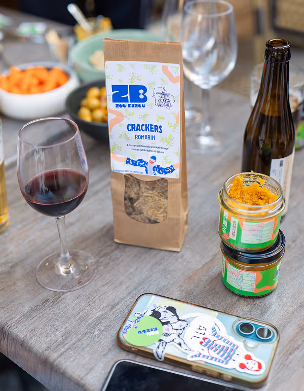 Paquet de crackers au romarin sur une table avec un verre de vin rouge, deux pots de tartinade, une bouteille de bière, et un téléphone avec des autocollant.