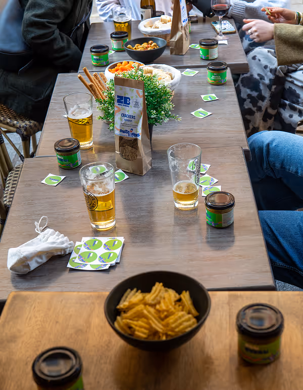 Table with snacks including chips, crackers, and olives, several glasses of beer and wine, green labeled jars, and people sitting around it.