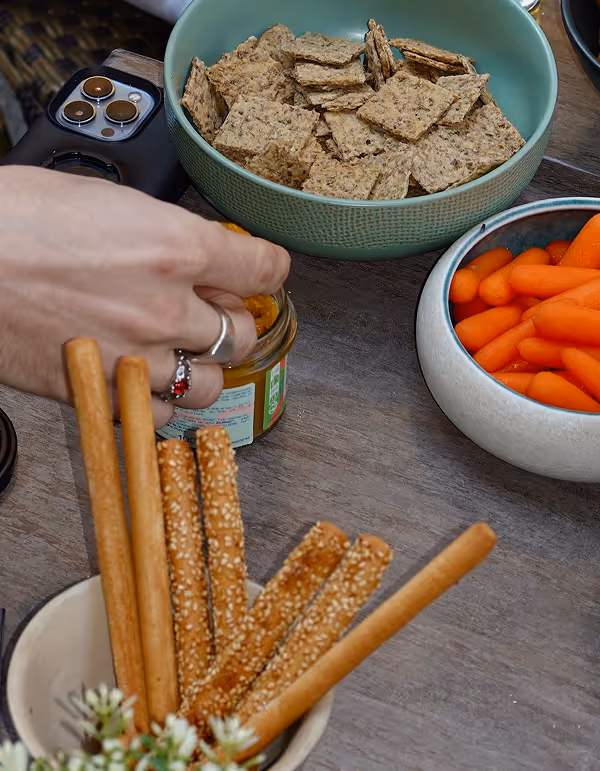 Main prenant un bâtonnet de snacks à tremper dans un pot de sauce, avec des bols contenant des crackers, des bâtonnets de pain et des mini-carottes sur une table en bois.