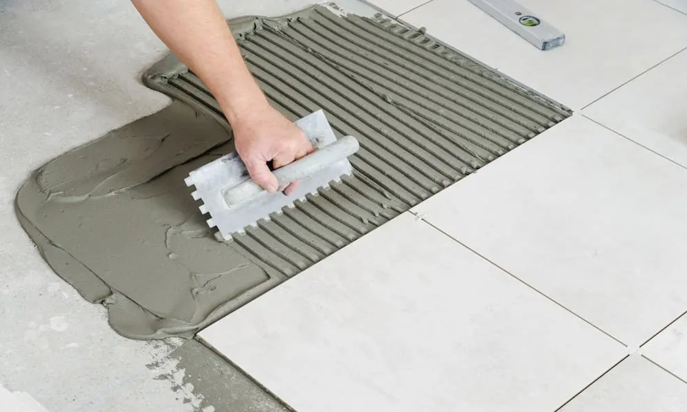 Fliesenleger kämmt Fliesenkleber mit der Zahnkelle auf und verlegt weiße Bodenfliesen im Verband