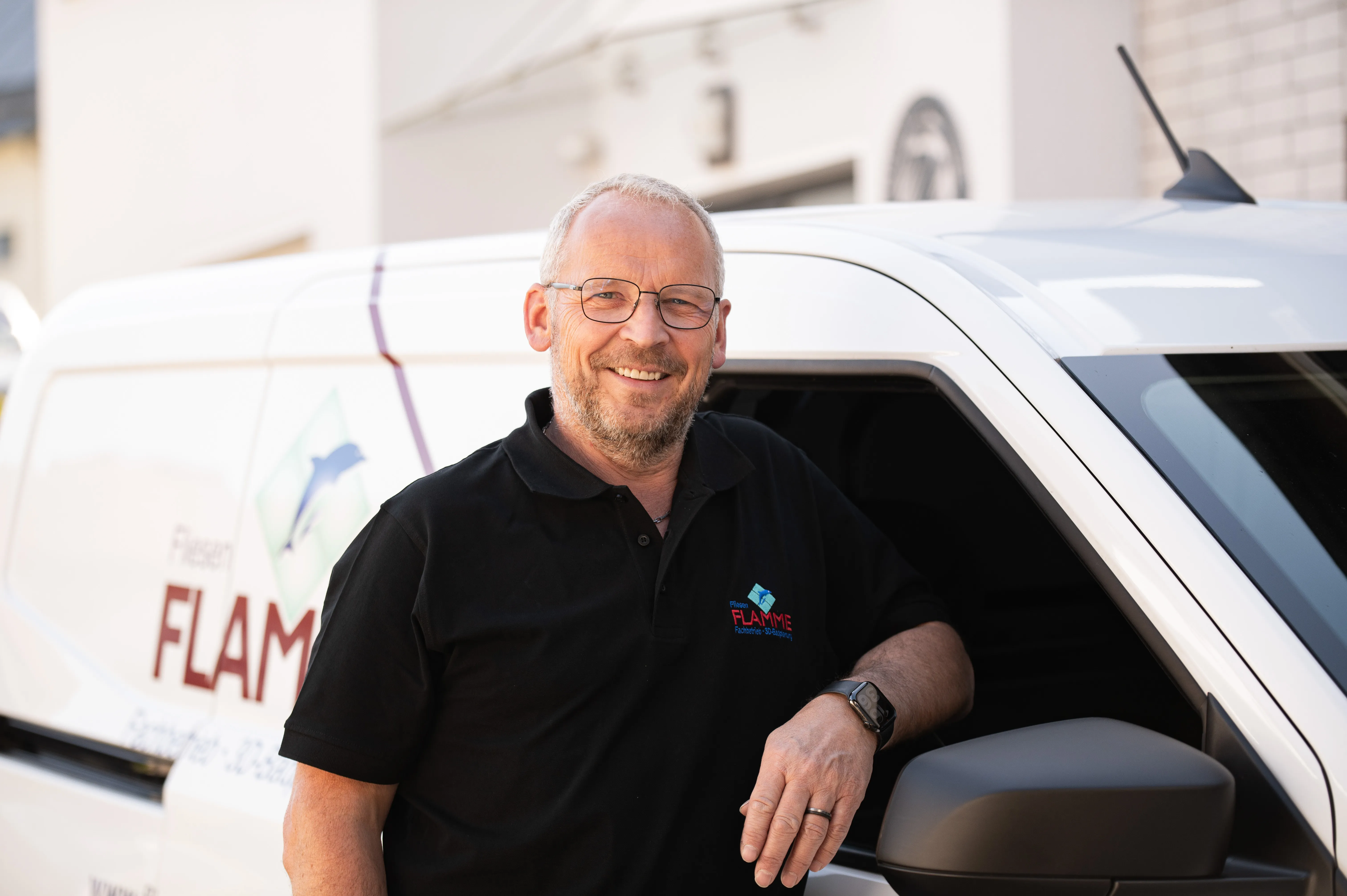 Lächelnder Mitarbeiter im schwarzen Polohemd mit Flamme-Logo vor dem Firmentransporter des Fliesenlegers