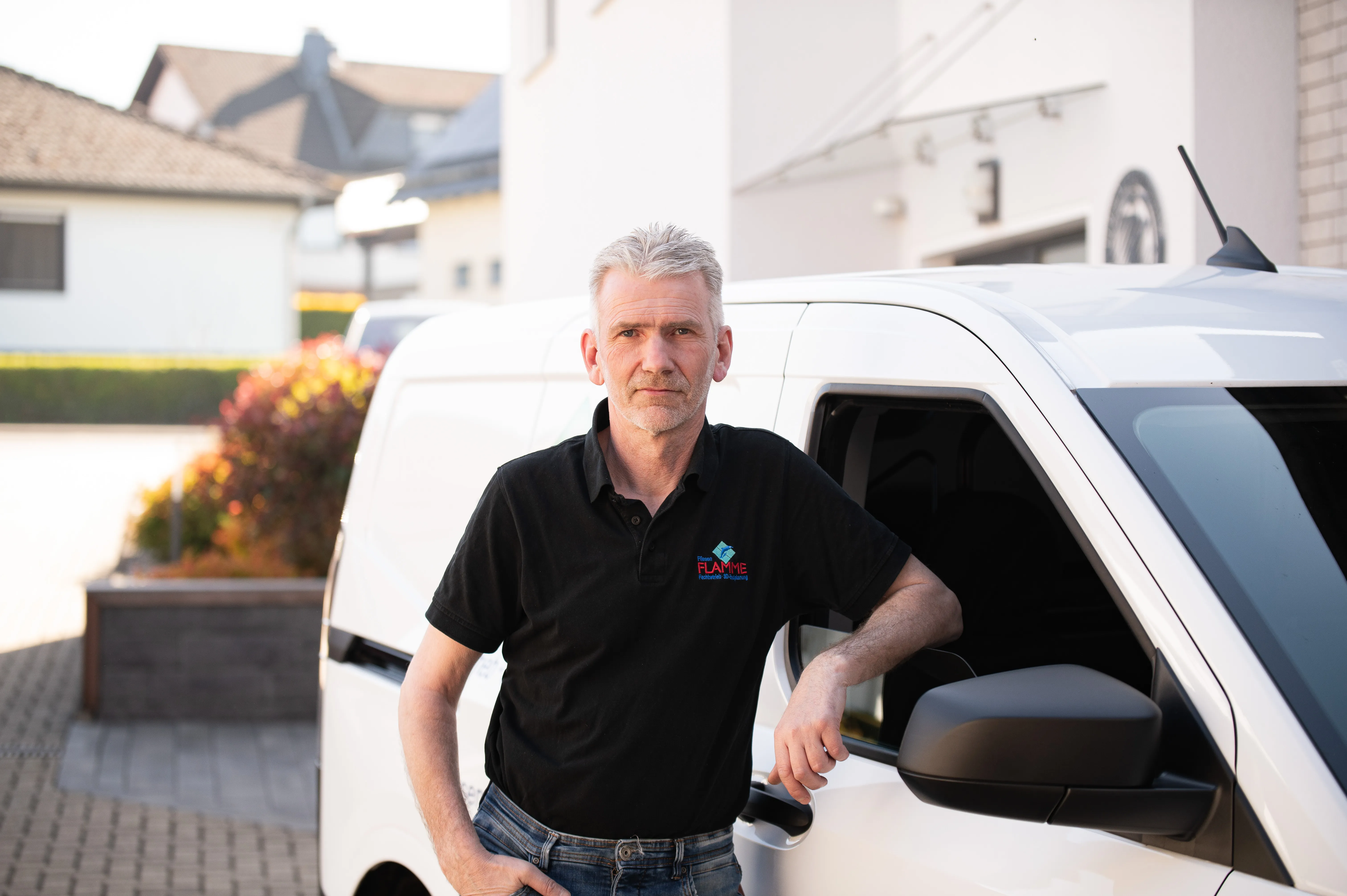 Fliesenleger im schwarzen Poloshirt mit Firmenlogo, angelehnt an den weißen Firmentransporter