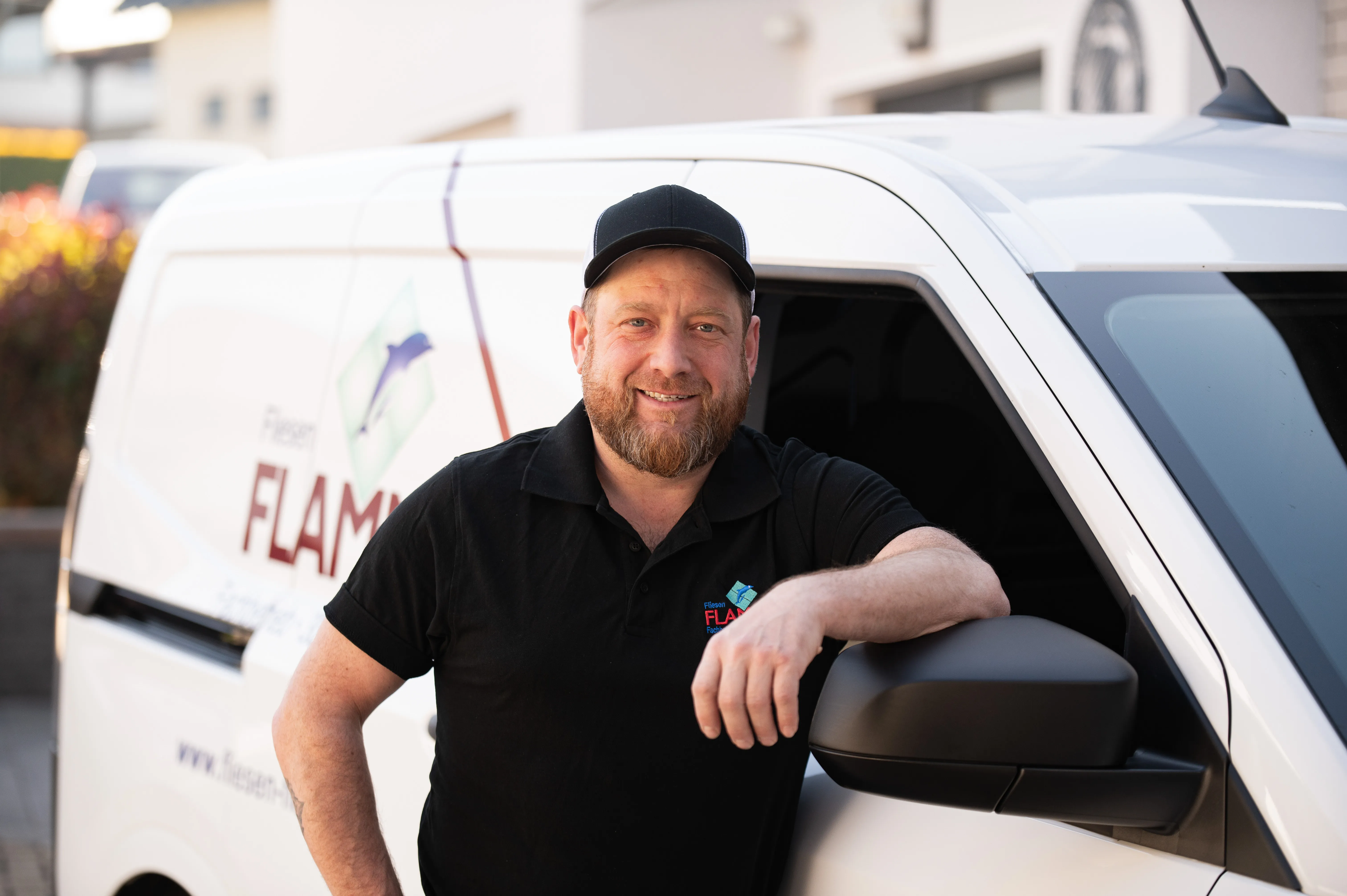 Fliesenleger mit Basecap und schwarzem Firmenpolo vor dem Transporter mit Flamme-Schriftzug