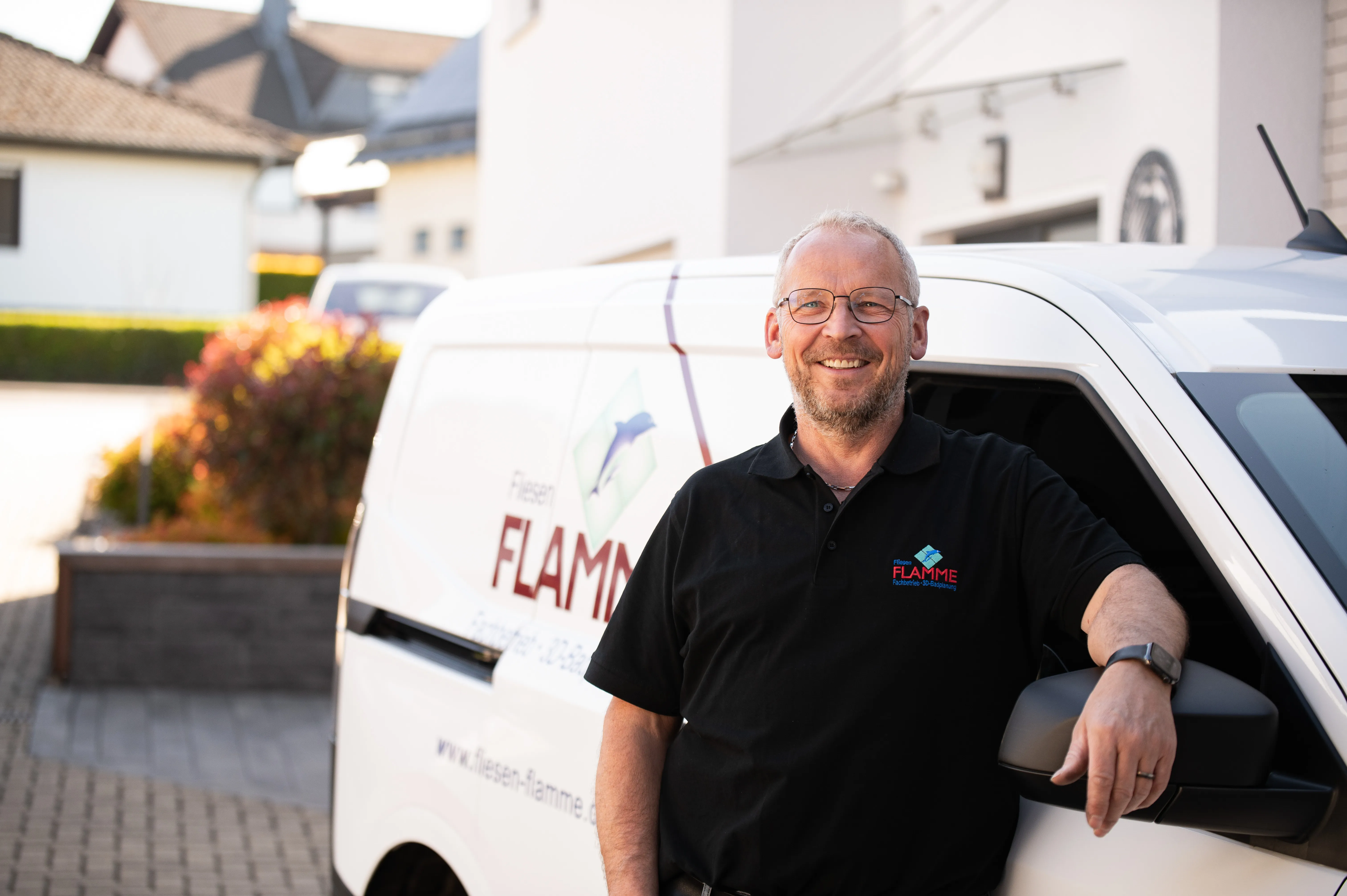 Fliesenleger mit Brille im schwarzen Firmenpolo vor dem Transporter mit Flamme-Schriftzug