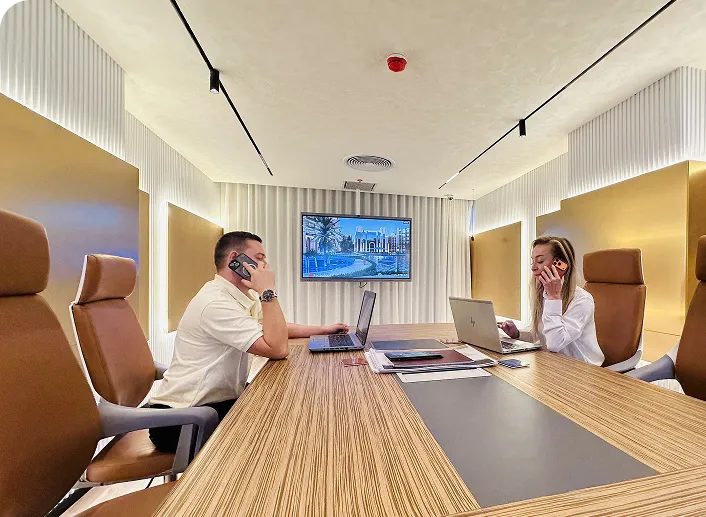 Two people sitting at a long conference table using laptops and talking on phones in a modern meeting room with a large screen on the wall.