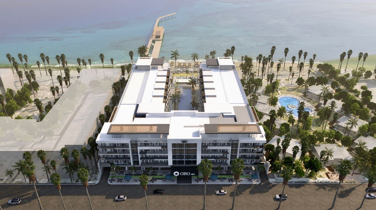 Aerial view of a modern U-shaped beachfront hotel with a central pool, palm trees, and oceanfront pier.