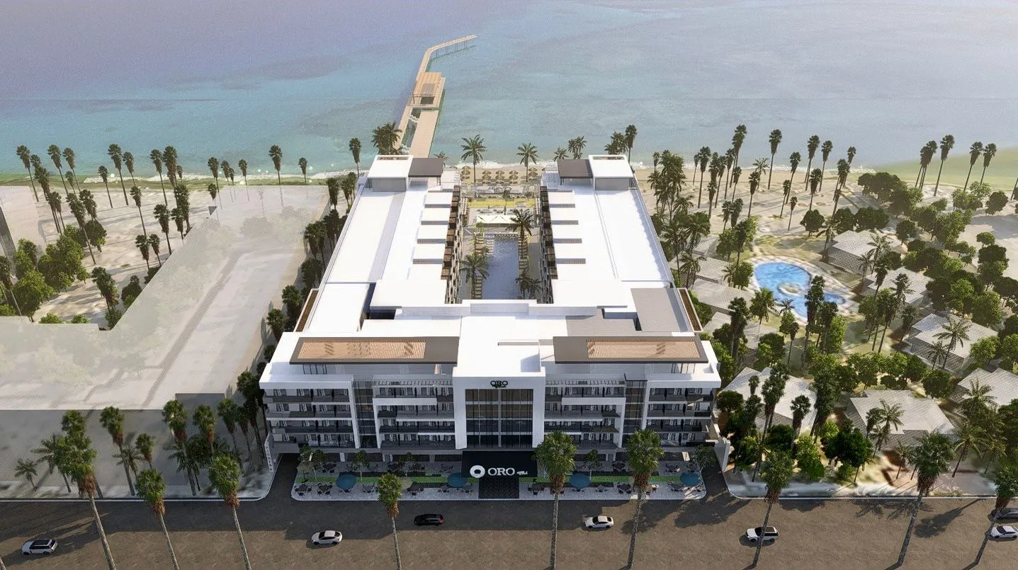 Aerial view of a modern U-shaped beachfront hotel with a central pool, palm trees, and oceanfront pier.