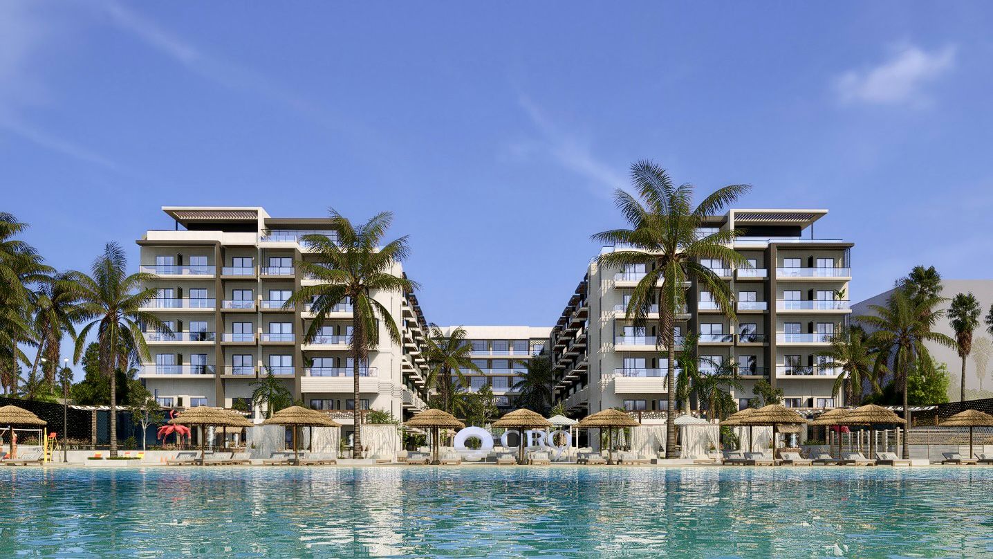 Modern resort buildings with balconies facing a large turquoise pool, surrounded by palm trees and thatched umbrellas under a clear blue sky.