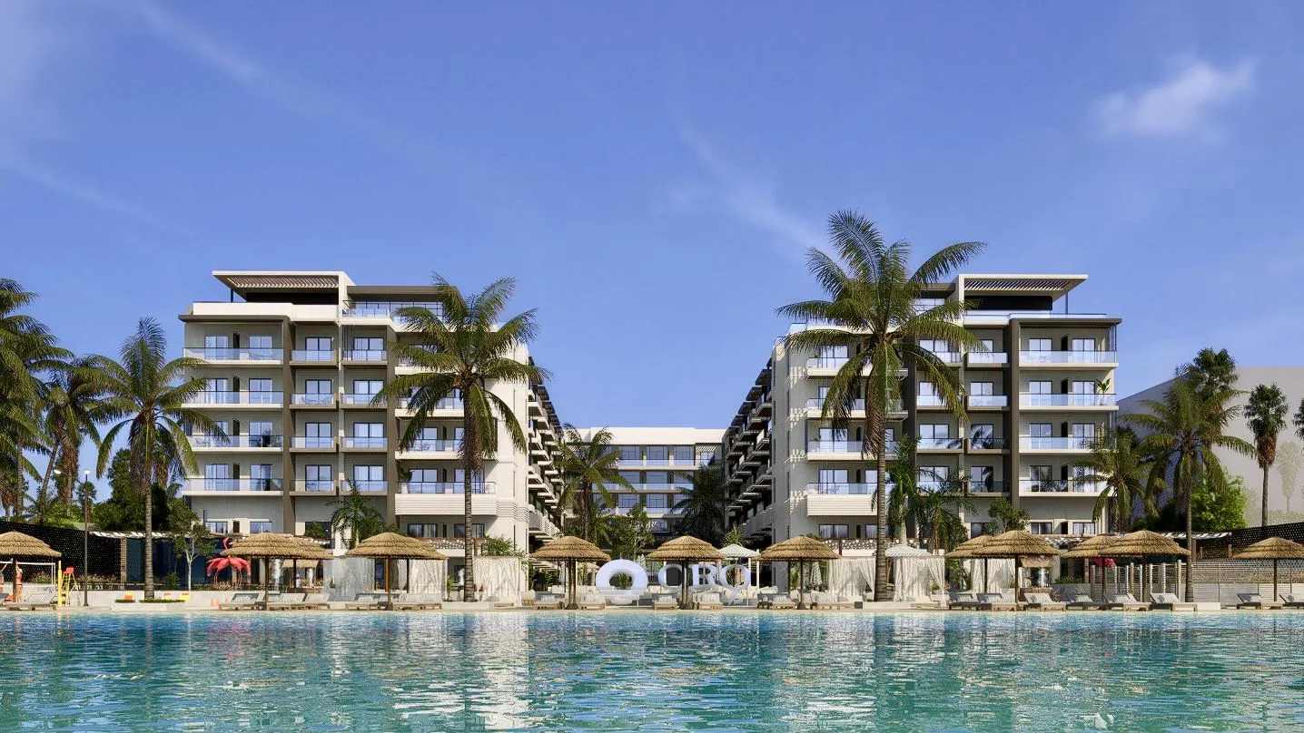 Modern resort buildings with balconies facing a large turquoise pool, surrounded by palm trees and thatched umbrellas under a clear blue sky.