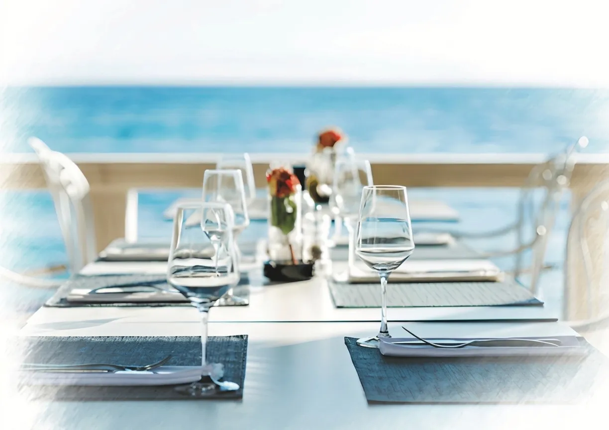 Dining table set with empty wine glasses and cutlery on blue placemats, overlooking a calm ocean view.
