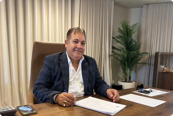 Man in a dark blazer sitting at a desk with papers and a pen, smiling at the camera.