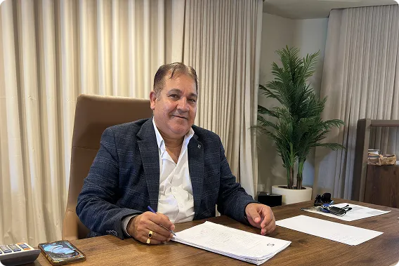 Man in a dark blazer sitting at a desk with papers and a pen, smiling at the camera.