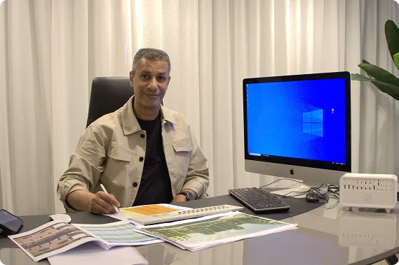 Man sitting at desk with documents and computer, holding a pen and smiling at the camera.