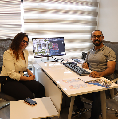 Two people sitting at a desk with architectural plans and a computer showing building designs.