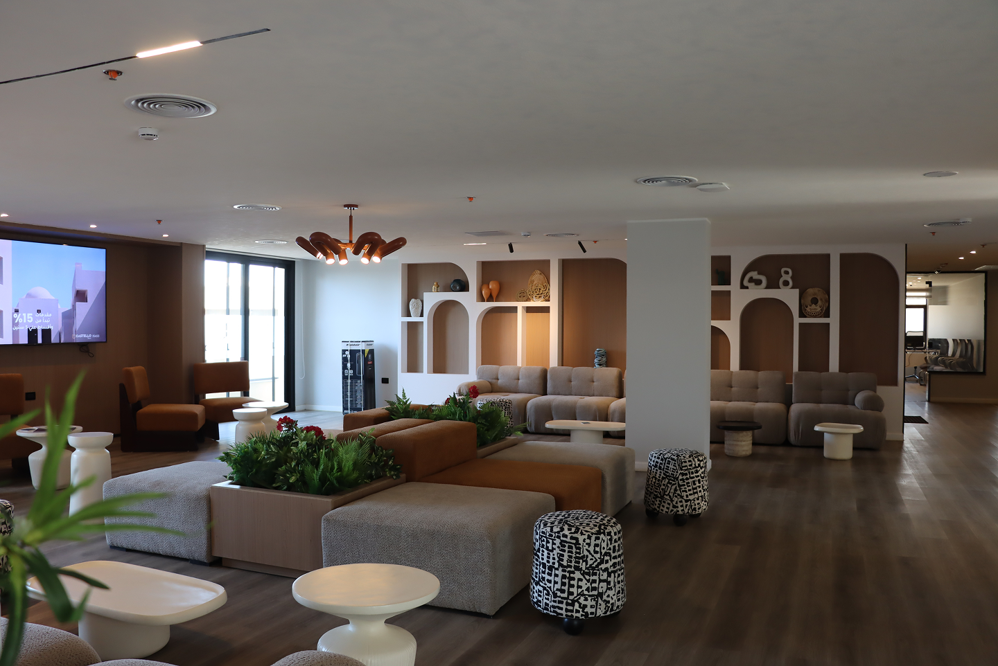Modern lounge with gray and brown sofas, plants, patterned stools, white coffee tables, and decorative shelves.