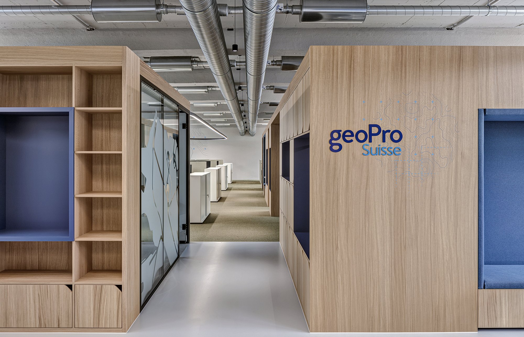 Moderner Bürogang mit Holzregalen, blauen Sitznischen und luftigen Deckenrohren, mit geoPro Suisse Logo an der Wand.