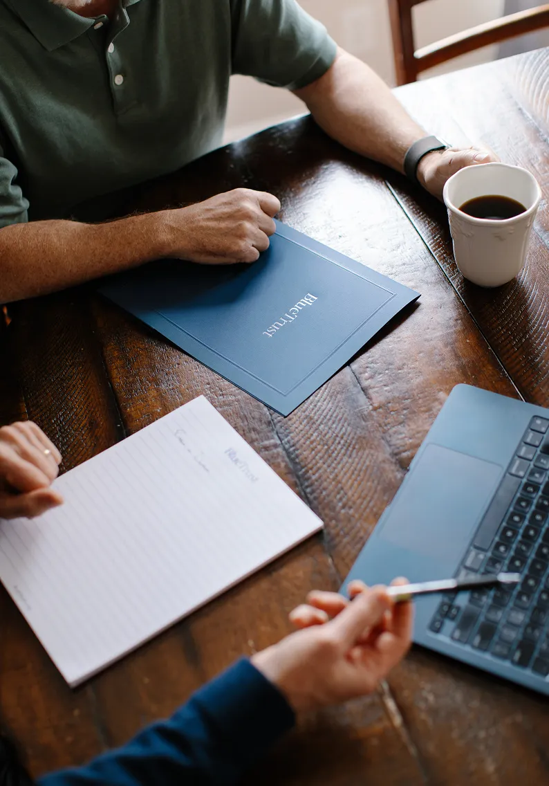 Two people sitting at a wooden table with a blue folder labeled 'BlueTrust,' a notepad, a laptop, and one person holding a pen while the other holds a cup of coffee.