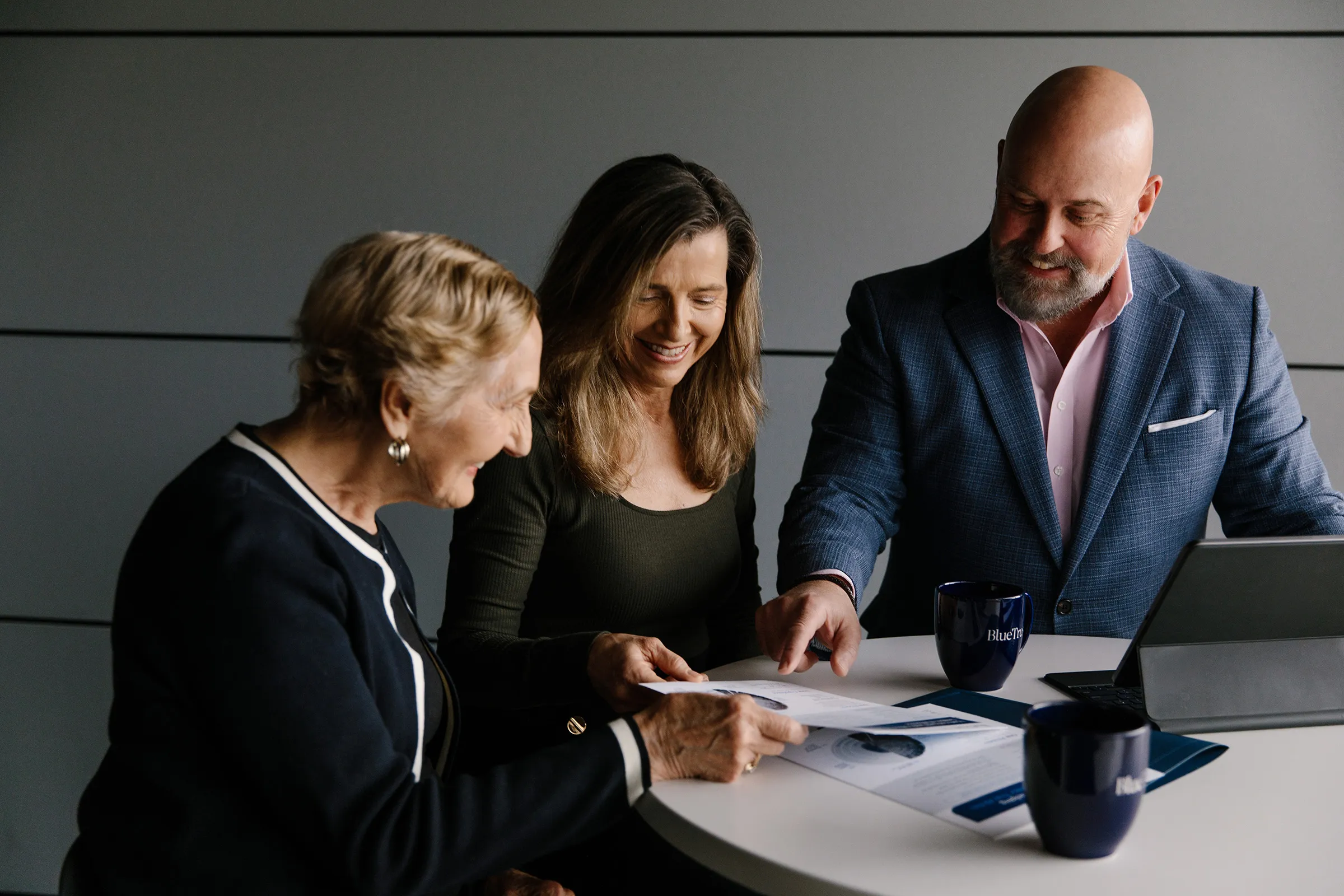 Three professionals gathered around a round table reviewing documents and smiling, with BlueTrust coffee mugs and a tablet.