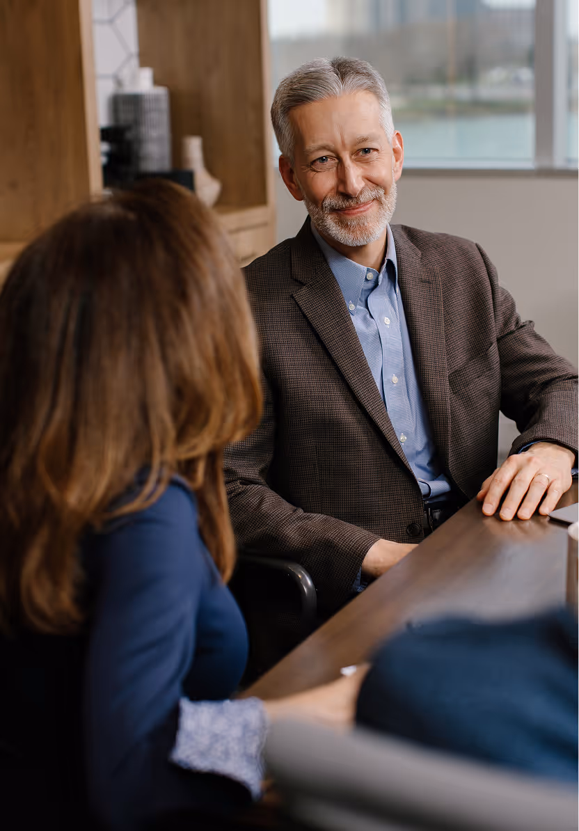 Smiling middle-aged man with gray hair in a brown blazer conversing with two people at a table in a modern office.