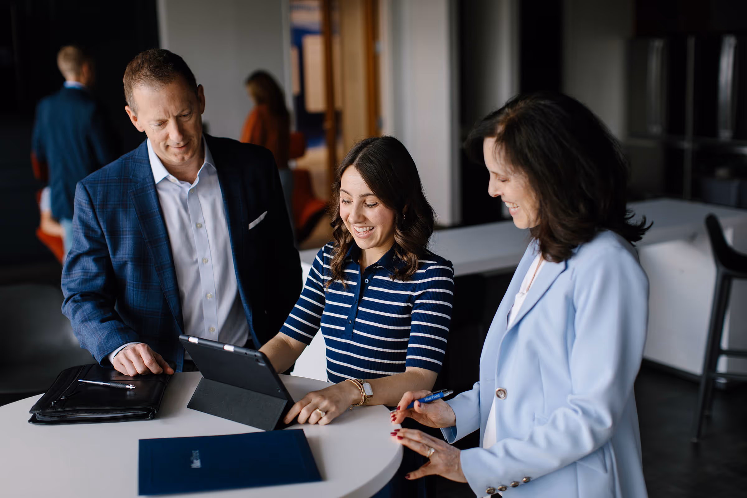 Three professionals gathered around a table looking at a tablet, smiling and engaged in discussion.