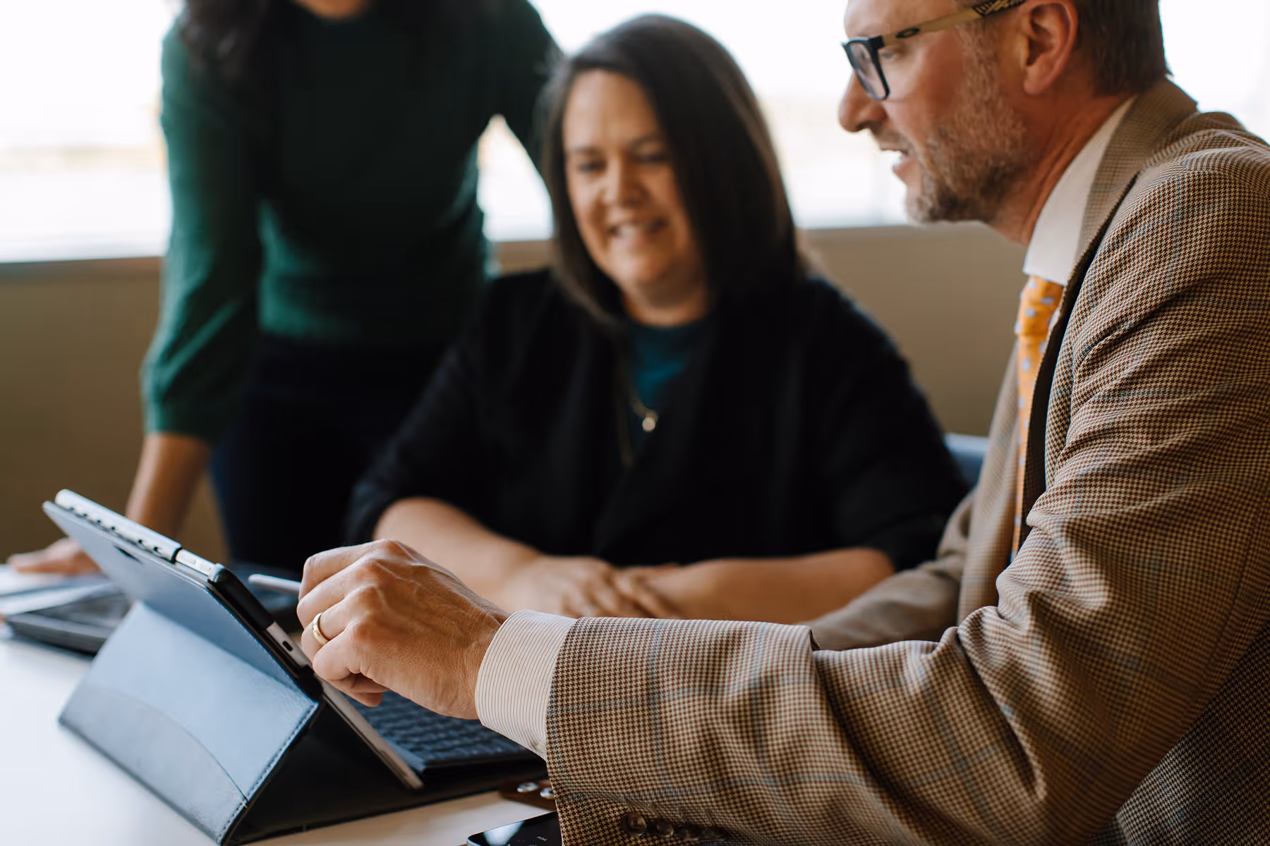 Three professionals gathered around a tablet at a desk, discussing work in a bright office.